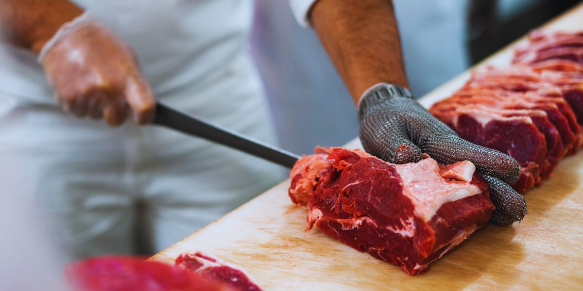 Mains d’un boucher qui découpe une pièce de viande rouge sur un billot de bois, couteau à désosser et ficelle de bridage à côté