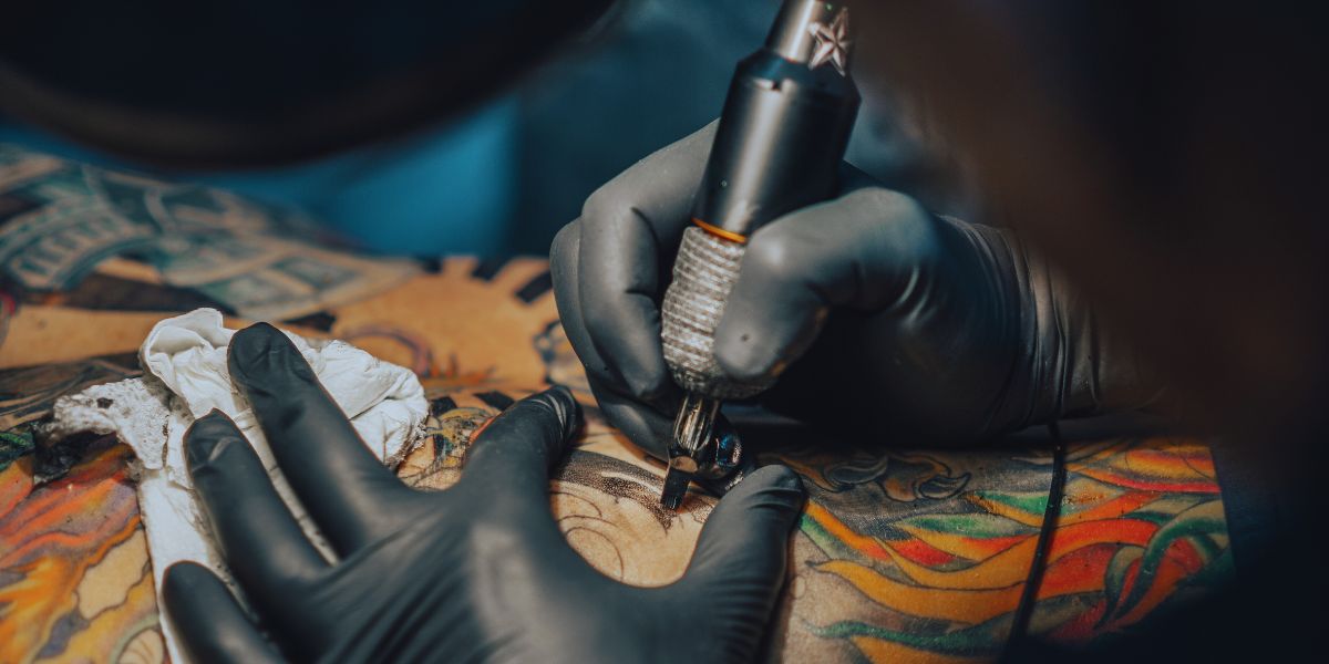 Main gantée d'un tatoueur tenant une machine à tatouer, aiguille posée sur un avant-bras aux lignes noires fraîches